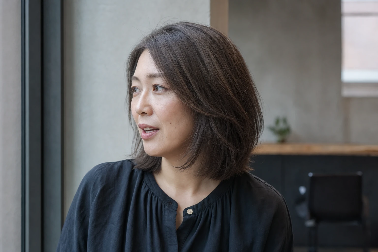 肩にかかるレイヤーボブのソフトアッシュブラウンを左三四分から自然光で見せる成人女性