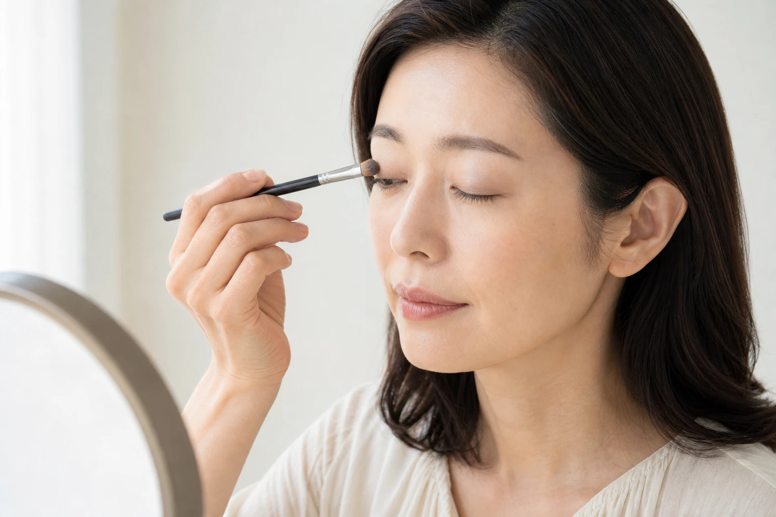 鏡の前で清潔な丸筆を右まぶたへ平行に当てて薄いブラウンを重ねる成人女性