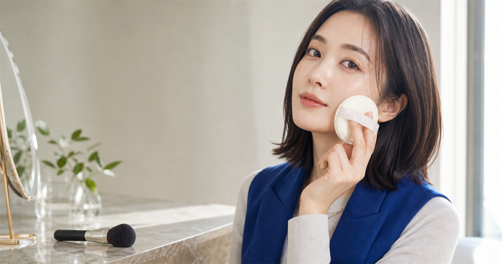 青いベストを着た女性がフェイスパウダーのパフを頬に当てる横長写真