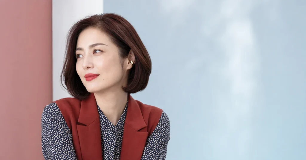 赤いリップと自然なグレーブラウン眉を合わせ、ブリックレッドのベストを着た成人女性